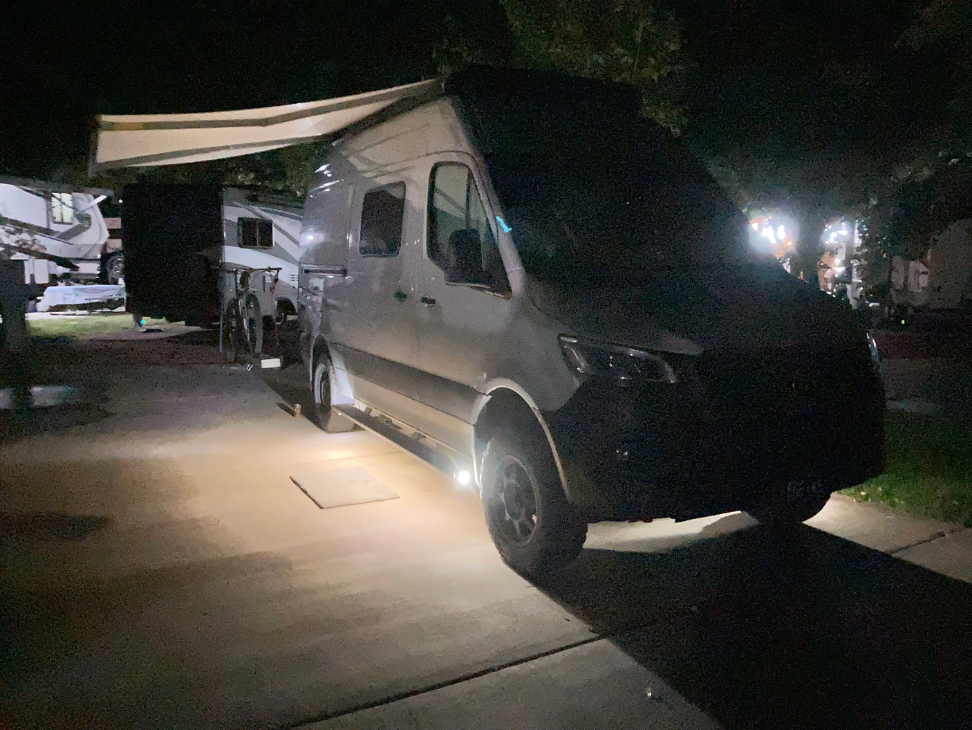 A white camper van, a common RV rental, is parked at a campsite at night, with an awning extended and under-vehicle lights on.