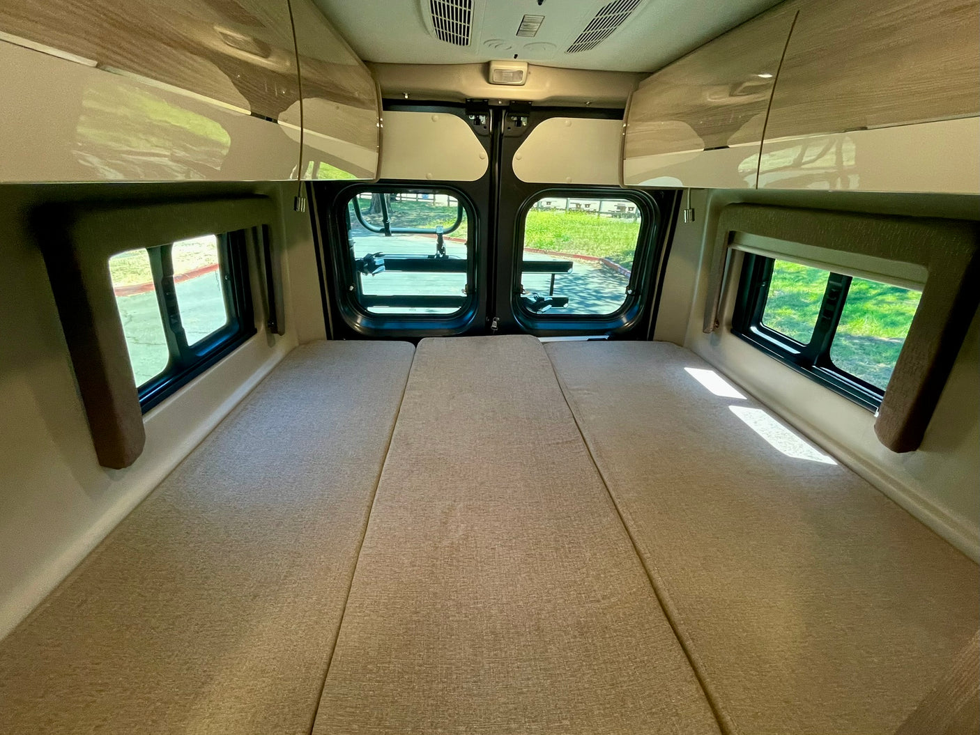 RV rental's rear interior: a bed or lounge area with light-colored cushions, large windows, and wood-toned overhead cabinets.
