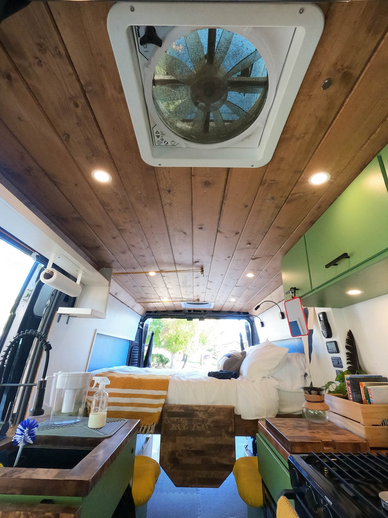 RV rental: A wide-angle view of a camper van interior with a wood-paneled ceiling, fan, bed, and kitchen area. — photo 17