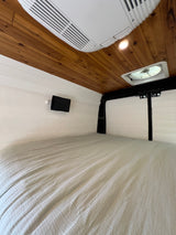Cozy RV rental interior with a comfortable bed, wood ceiling, white walls, AC, fan, and a mounted tablet.