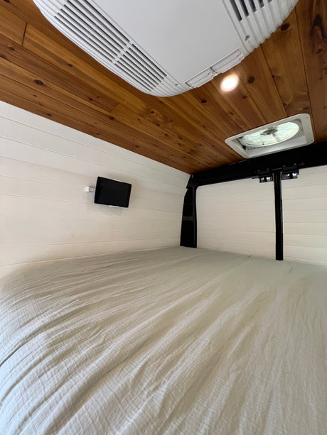 Cozy RV rental interior with a comfortable bed, wood ceiling, white walls, AC, fan, and a mounted tablet.
