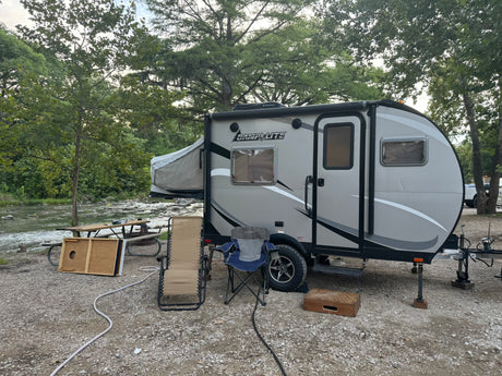 RV rental campsite by a river: a grey camper with a pop-out bed, picnic table, and chairs on gravel.