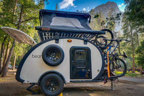 This sleek teardrop RV rental, featuring a rooftop tent and bikes, is set in a scenic forest campground with a mountain view.