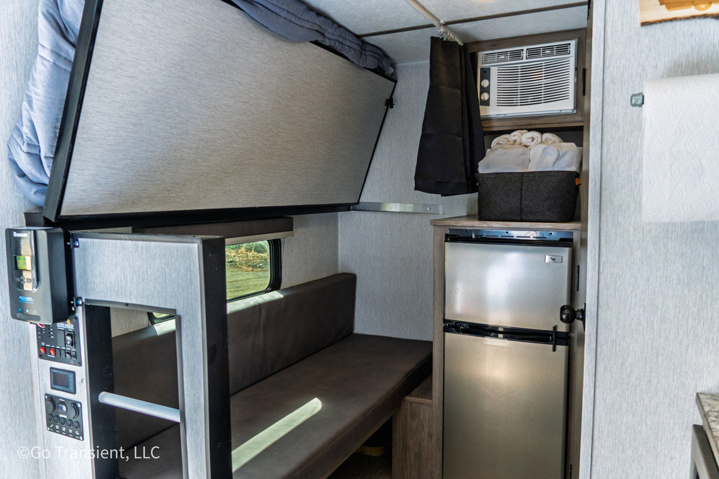 Compact RV rental interior featuring a fold-down bunk, sofa, small fridge, and AC unit. Ideal for travel.