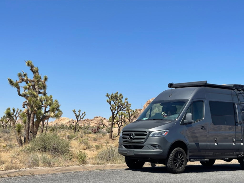 A grey Mercedes Sprinter RV rental van is parked amidst Joshua Trees in a sunny desert landscape.