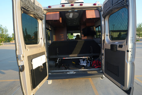 View into an RV rental from the open rear doors, showing the bed, overhead cabinets, TV, and stored items.