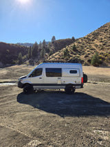 Explore nature in this adventure-ready RV rental! Silver camper van on a sunny dirt road by dry hillsides & a stream.