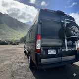 A dark gray camper van RV rental sits on a gravel lot, backed by misty green mountains and clouds under a blue sky.