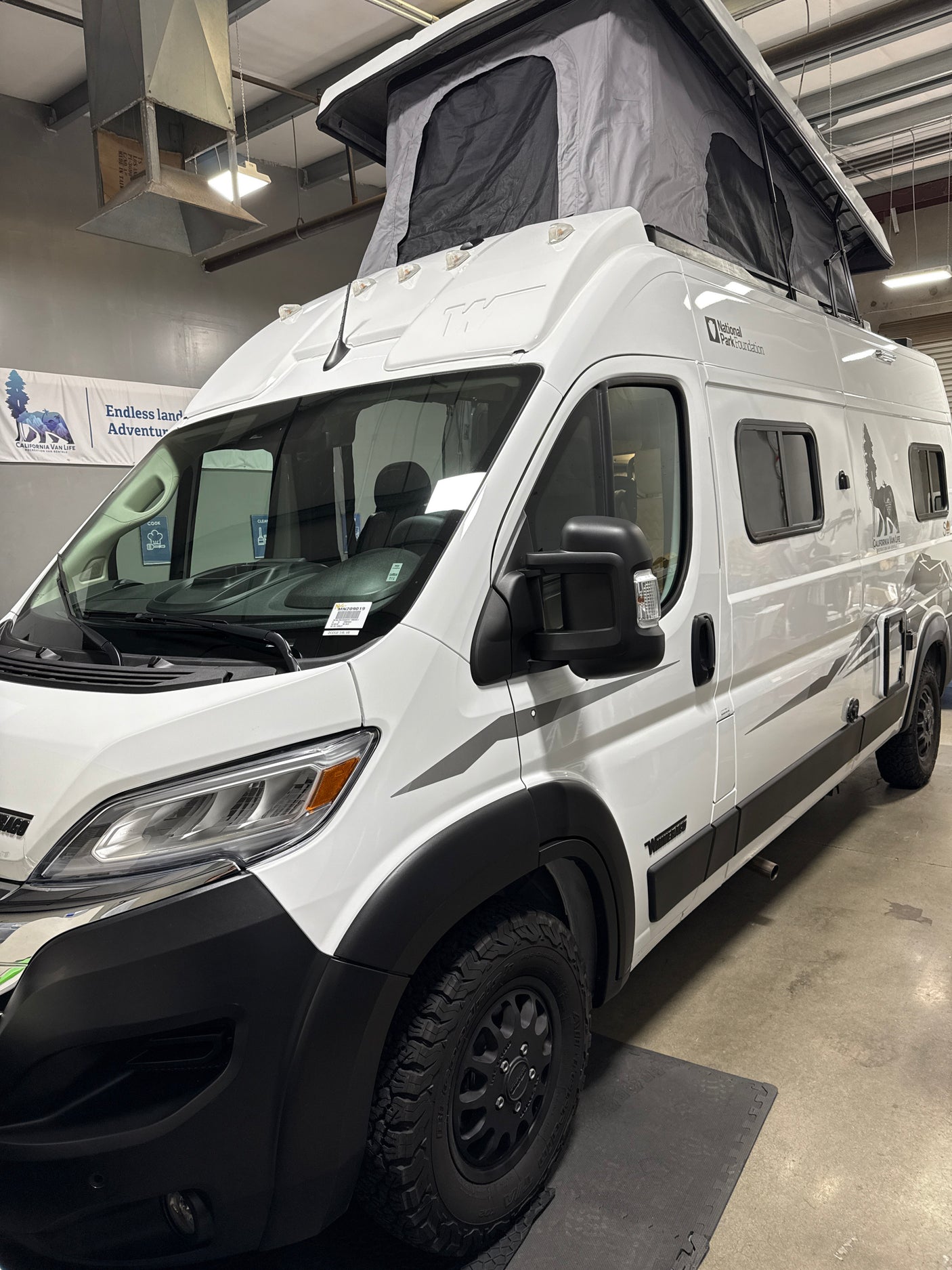 White camper van with pop-top tent, branded for California Van Life RV rental, parked indoors.
