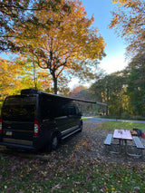 Black RV rental parked in an autumn campsite with extended awning, picnic table, and fire pit under colorful trees.