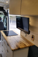 Inside a sleek RV rental van: a kitchen with wood counter, induction cooktop, drawers, and mounted TV. Compact living ready.