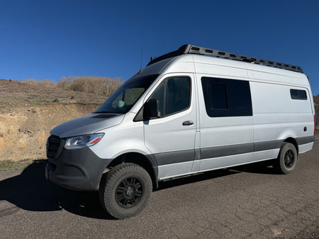Silver Sprinter camper van with off-road tires & roof rack parked on an asphalt road against dry hills & blue sky. Ideal rv rental.