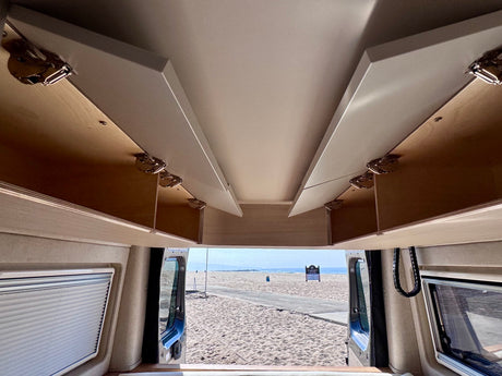 This cozy RV rental features convenient overhead storage and open rear doors revealing a sandy beach and ocean view.