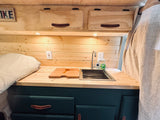 Cozy RV rental kitchenette features wood paneling, sink, custom cabinets, and nearby sleeping area.