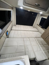 Spacious RV rental interior featuring light-colored cushions arranged as a bed, with windows & control panel access.