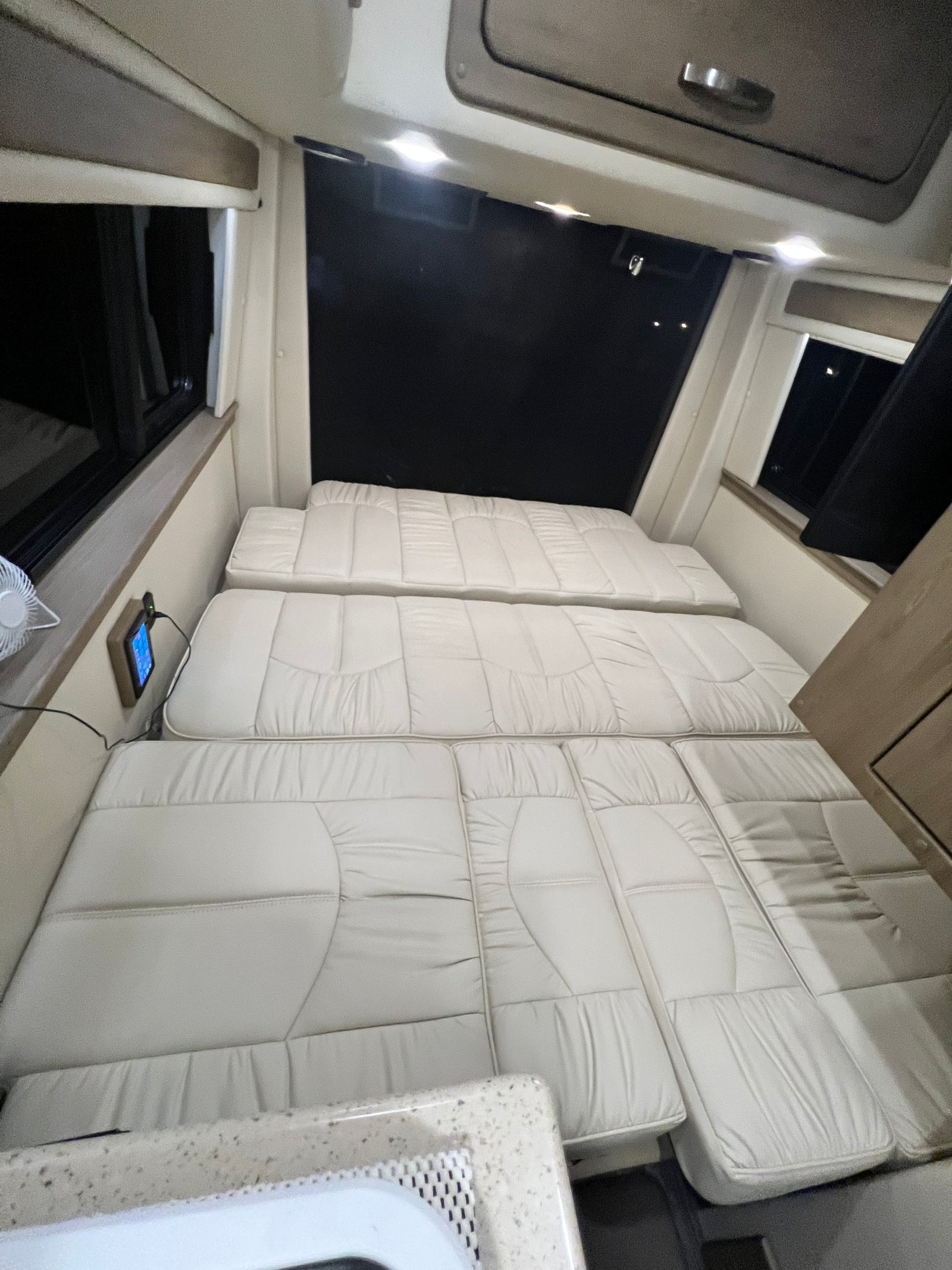 Spacious RV rental interior featuring light-colored cushions arranged as a bed, with windows & control panel access.