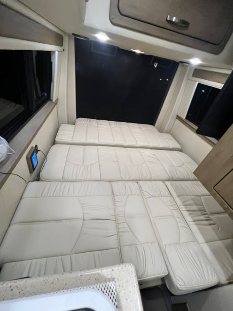 Spacious RV rental interior featuring light-colored cushions arranged as a bed, with windows & control panel access.
