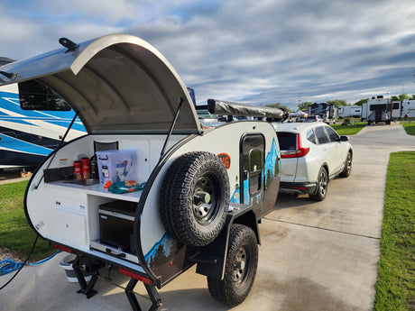 Compact rv rental teardrop camper with outdoor kitchen set up, hitched to SUV at an RV park.