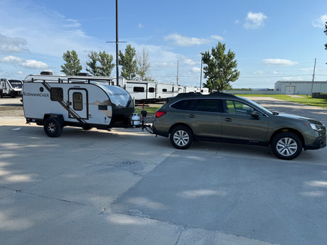 A dark green Subaru Outback tows a compact Bushwhacker rv rental camping trailer in a sunny outdoor lot with other RVs.