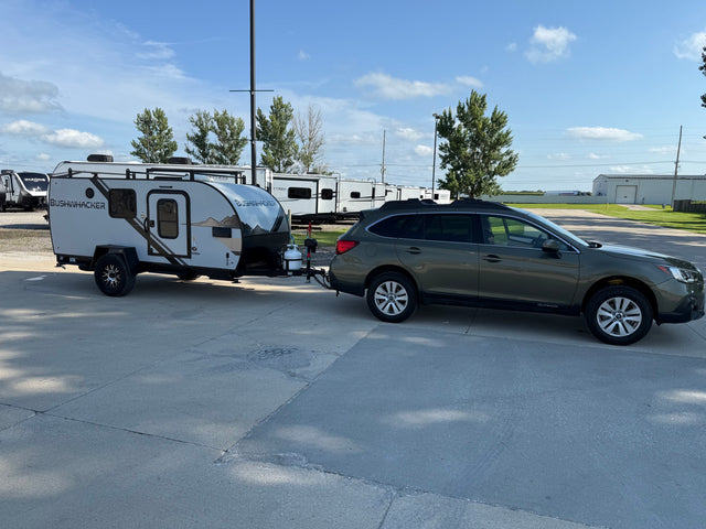 A dark green Subaru Outback tows a compact Bushwhacker rv rental camping trailer in a sunny outdoor lot with other RVs.
