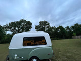 Compact, retro RV rental trailer with white & green exterior, parked in a grassy field under an overcast sky.