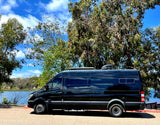 This black RV rental van, parked by a calm lake with trees and a blue sky, offers outdoor adventure.