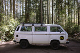 White 4WD VW Vanagon RV rental with roof rack, parked in a lush forest for adventure.