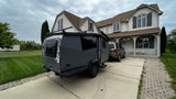 A gray Cricket camper, perfect for an RV rental, sits in a driveway next to an SUV at a large house.