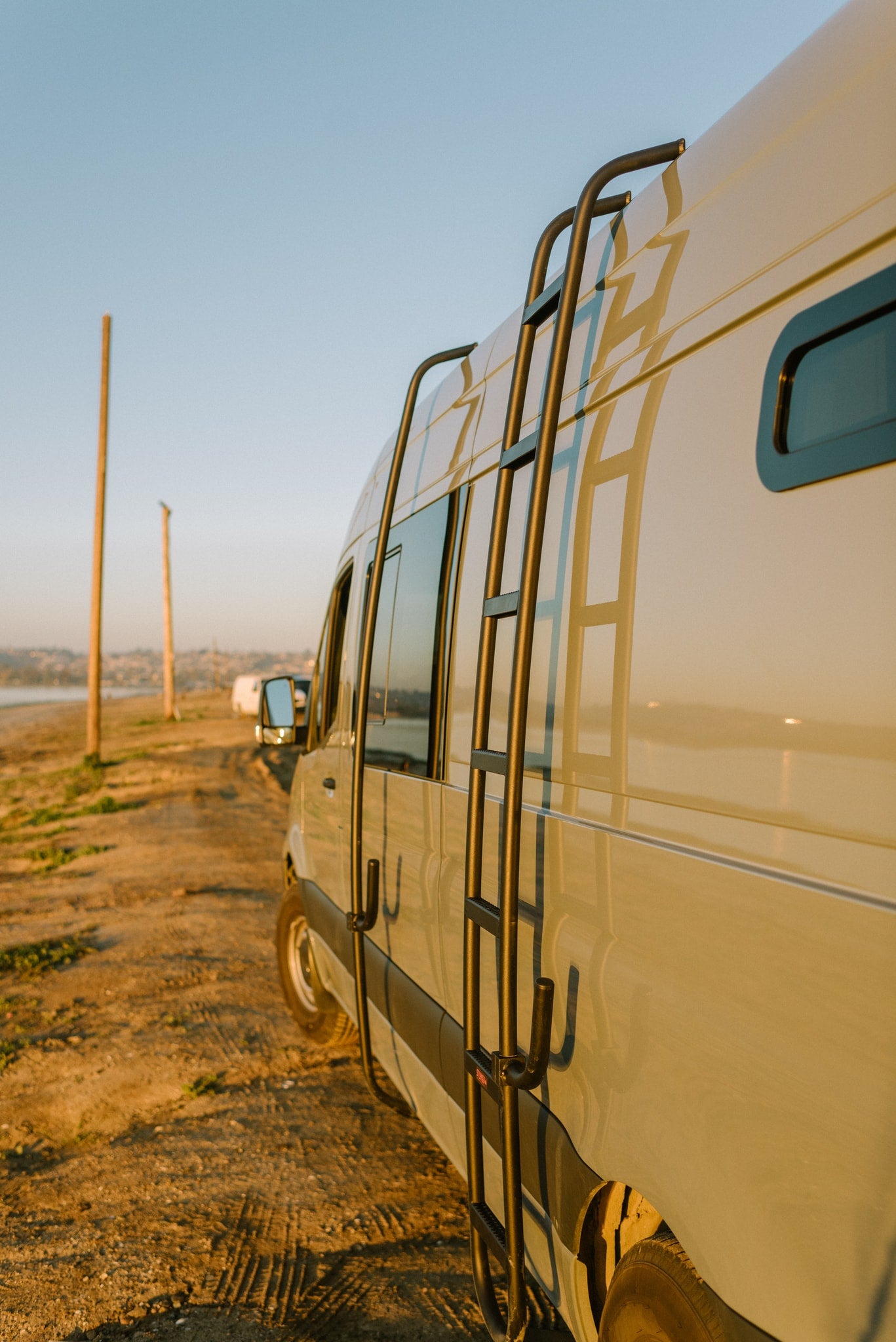 Golden hour over a light-colored RV rental van with a side ladder, parked on a dirt road by water. — photo 16