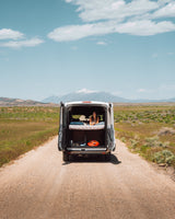 A woman relaxes in an RV rental van's open back, enjoying mountain views from her bed on a remote dirt road.