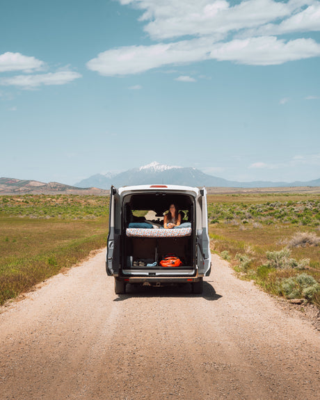 A woman relaxes in an RV rental van's open back, enjoying mountain views from her bed on a remote dirt road.