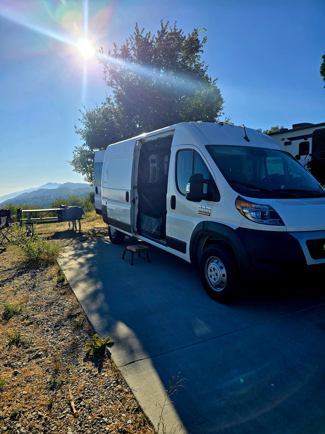 White rv rental van at a mountain campsite, door open, picnic table & views under a bright sun. Ideal for travel.