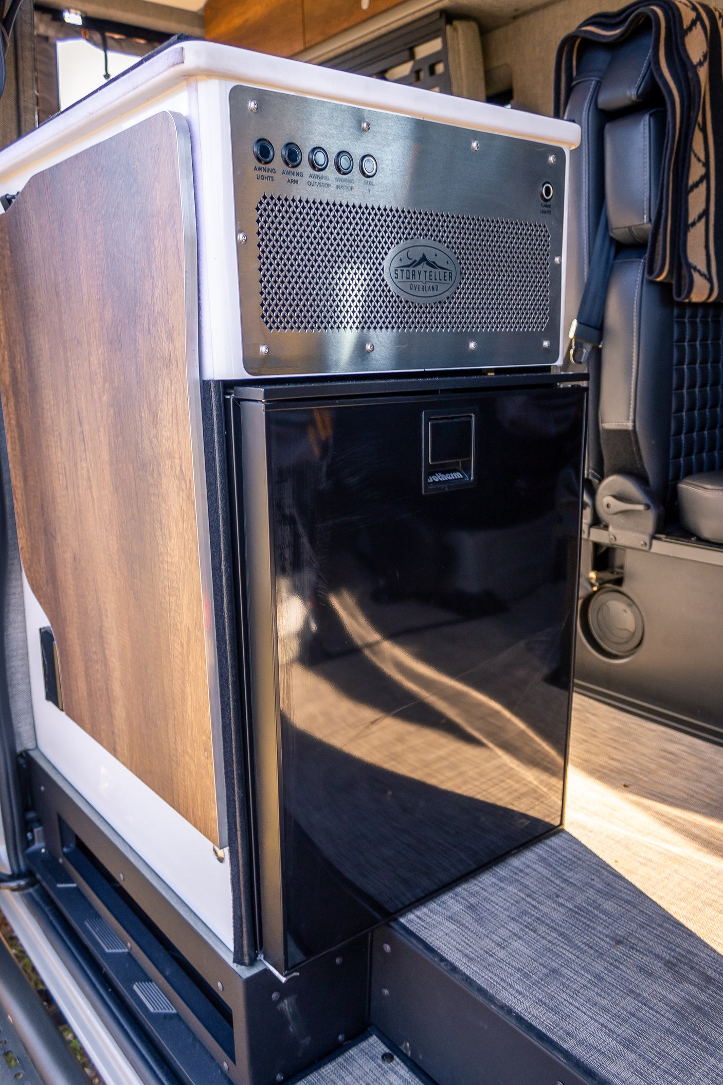 This rv rental interior features a black Webasto fridge beneath a metallic control panel with awning/light controls, next to a wood-paneled cabinet.
