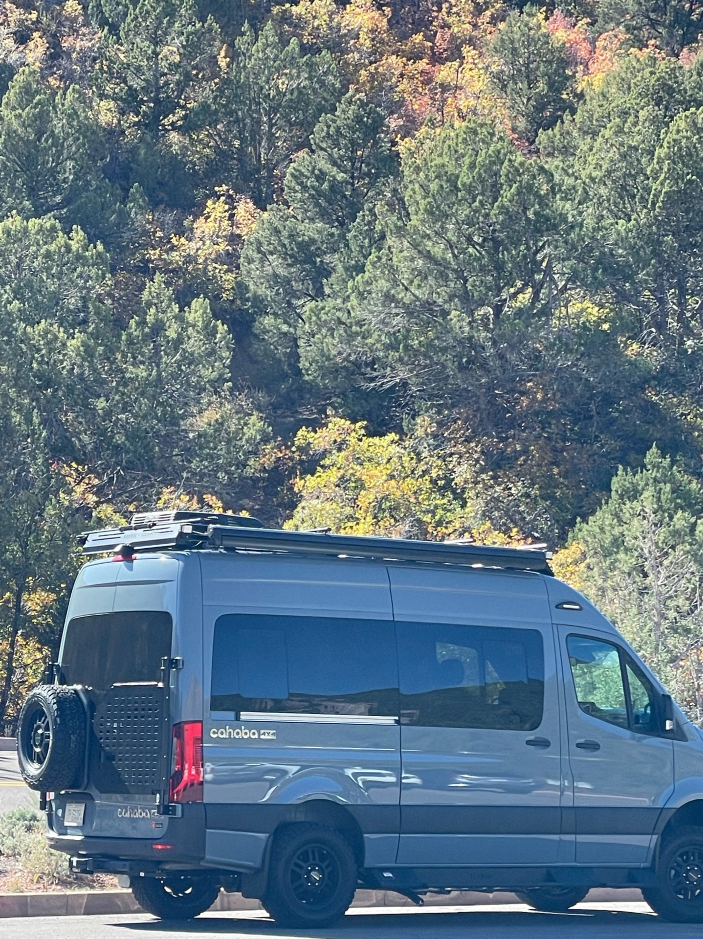 A gray Cahaba 4x4 Mercedes Sprinter camper van, ideal for RV rental adventures, parked by fall foliage.