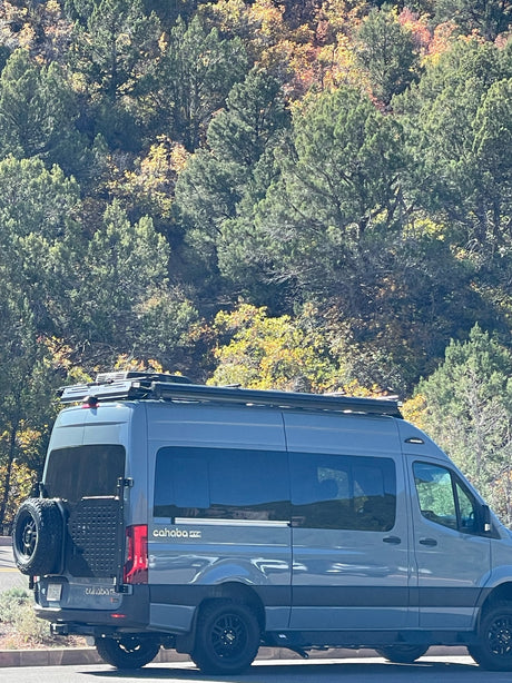 A gray Cahaba 4x4 Mercedes Sprinter camper van, ideal for RV rental adventures, parked by fall foliage.