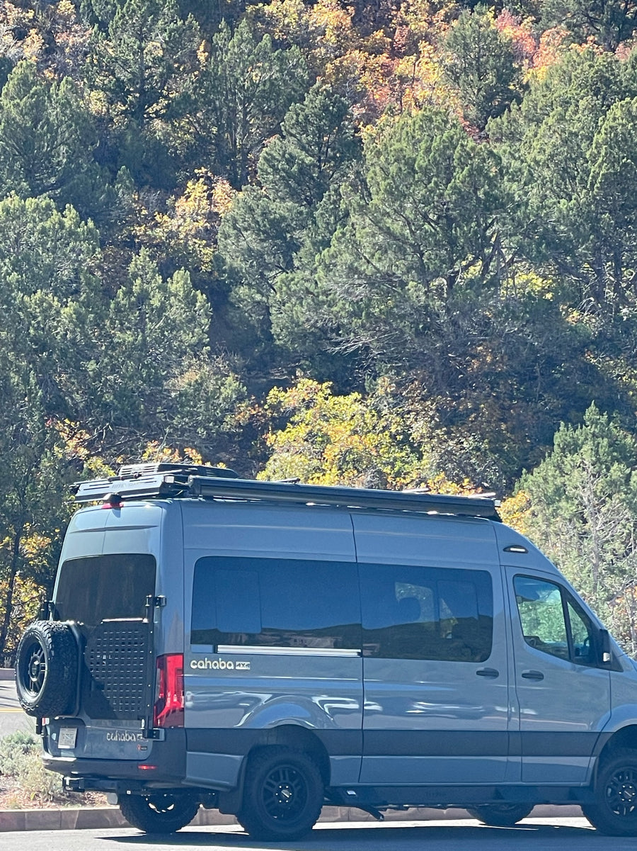 A gray Cahaba 4x4 Mercedes Sprinter camper van, ideal for RV rental adventures, parked by fall foliage.