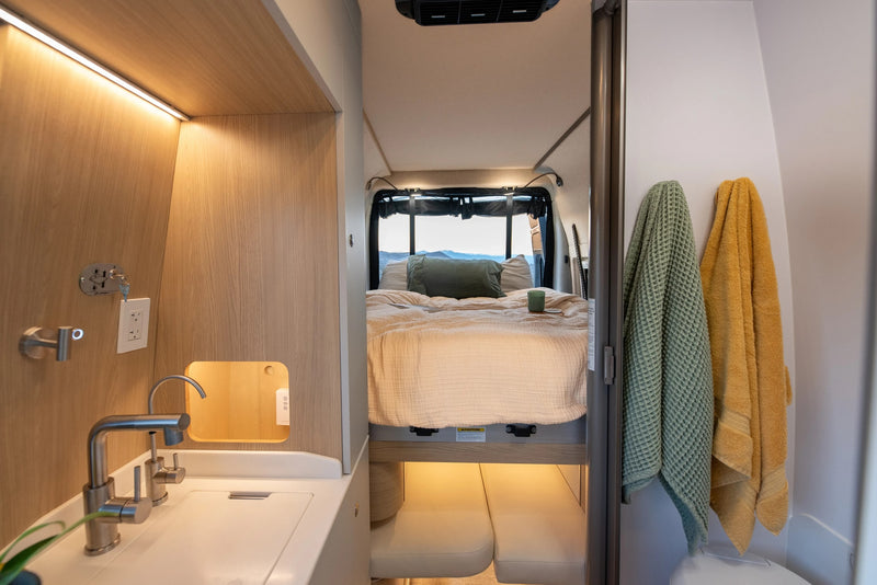 RV rental showing a compact kitchen with sink, a cozy bed with pillows, and scenic mountain views from the rear. — photo 7
