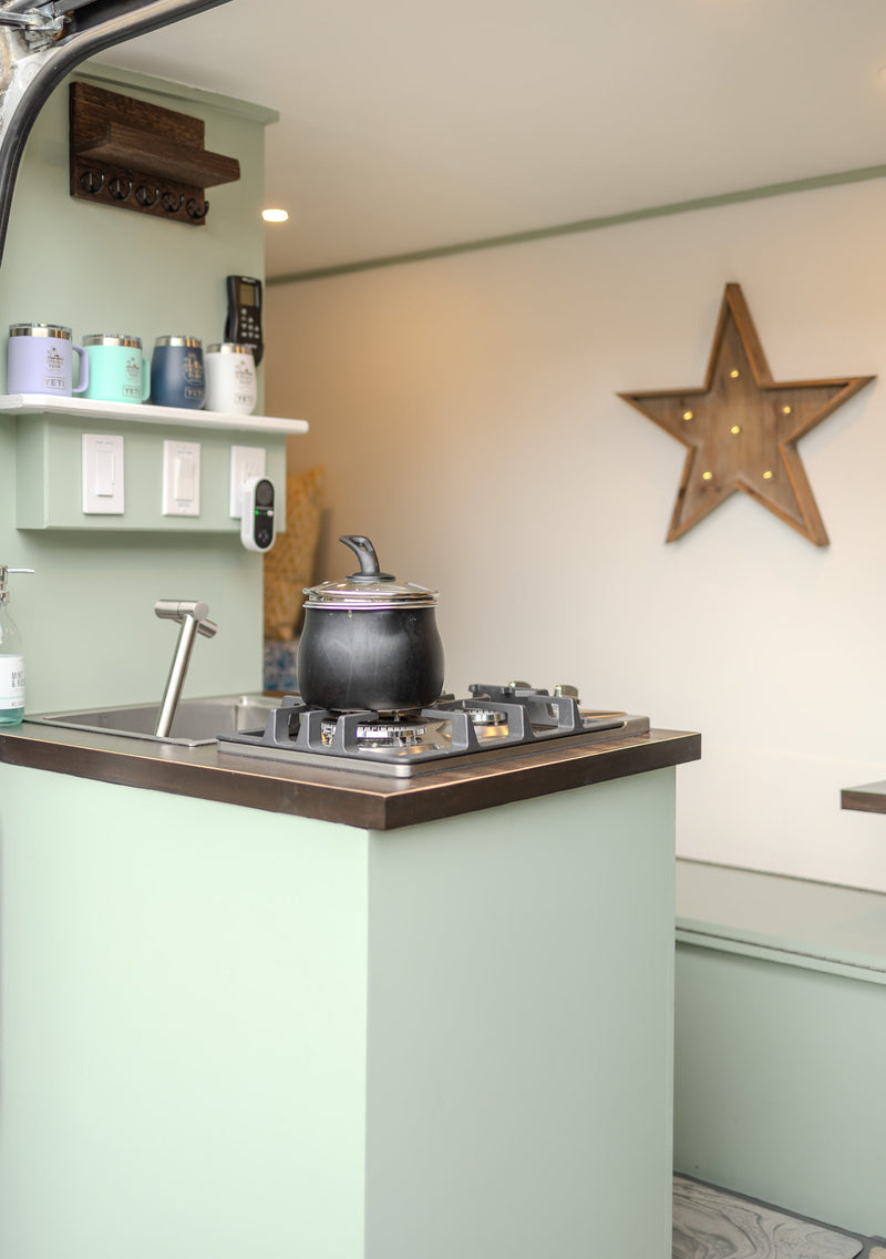 A bright, light green RV rental kitchen with a small stove, sink, and shelf of colorful mugs, designed for efficient travel living. — photo 7
