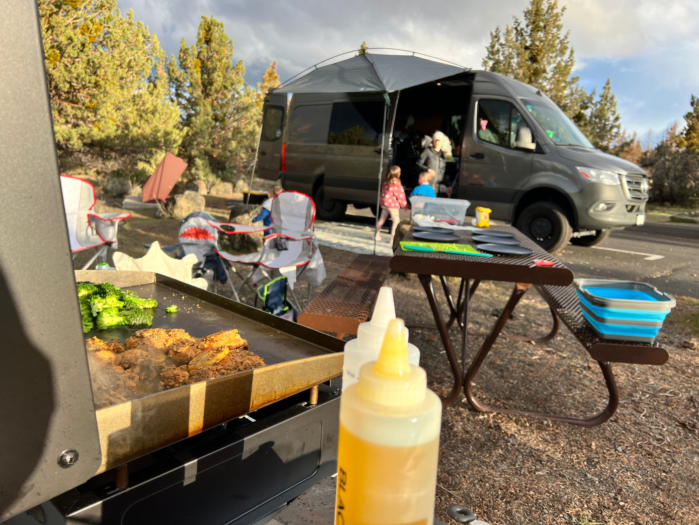 Family camping with an RV rental, cooking dinner on a griddle with broccoli & meat. Van, picnic table, chairs, and kids visible.