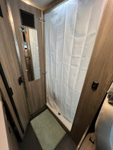 An rv rental's compact bathroom, featuring a shower with curtain, mirror, and light green floor mat.