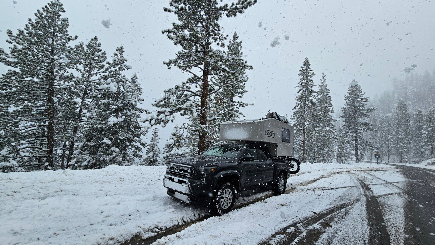 A rugged truck camper, ideal for an RV rental, is parked on a snowy forest road amidst tall pine trees.