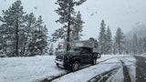 A rugged truck camper, ideal for an RV rental, is parked on a snowy forest road amidst tall pine trees.