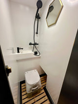 Modern RV rental bathroom with black shower, sink, Dometic toilet, wood slat floor, and hexagonal mirror.
