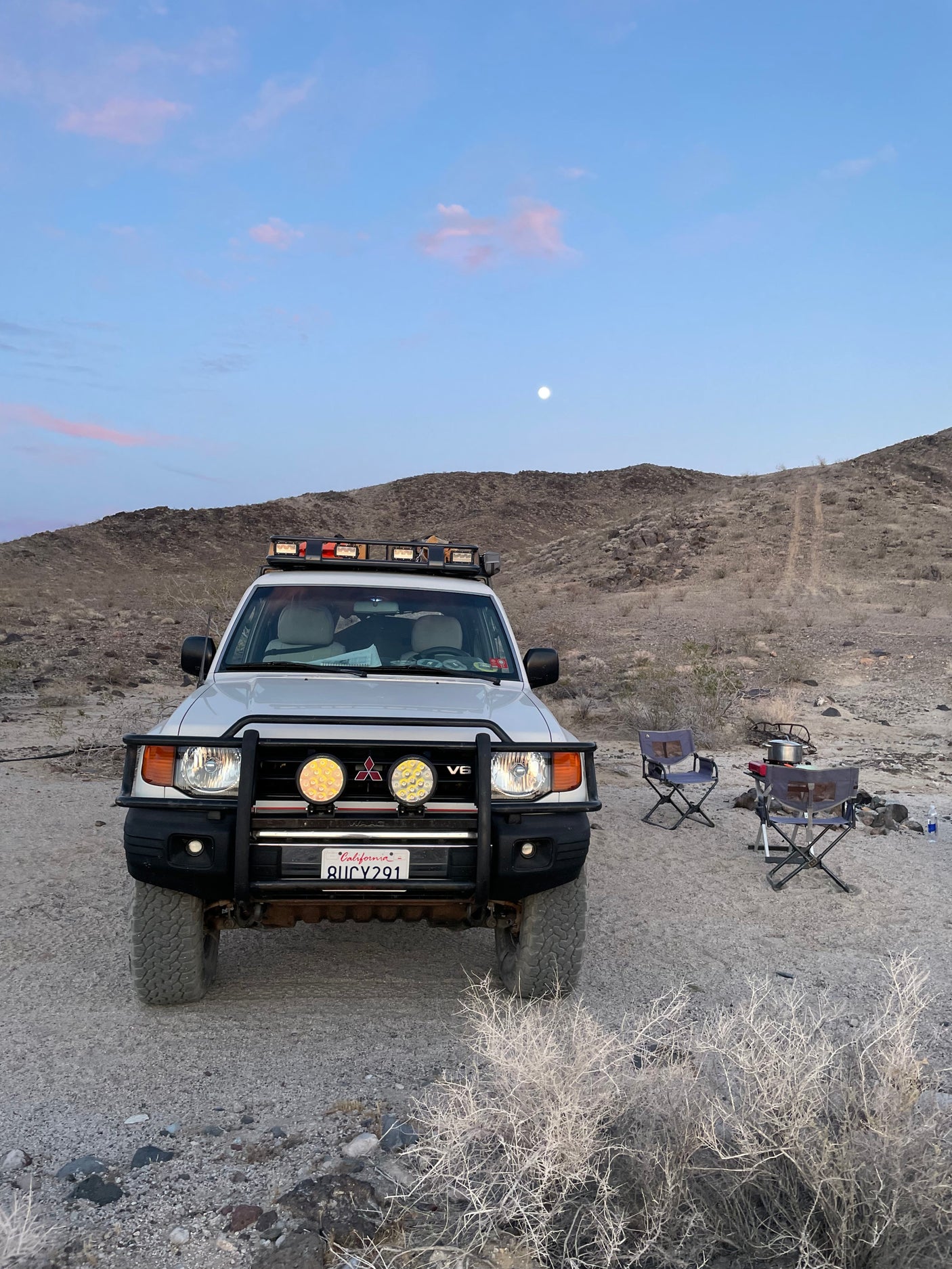 Moonlit desert camp with a white off-road SUV, serving as a functional RV rental. Chairs, stove & gear set up for adventure.