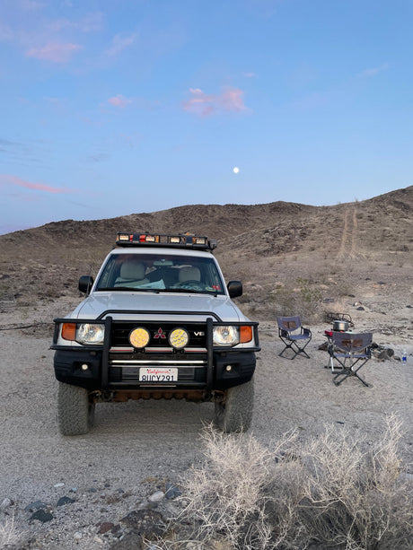 Moonlit desert camp with a white off-road SUV, serving as a functional RV rental. Chairs, stove & gear set up for adventure.