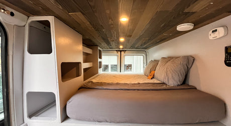 Inside a cozy RV rental: a well-appointed camper van interior with bed, built-in storage, & wood ceiling.