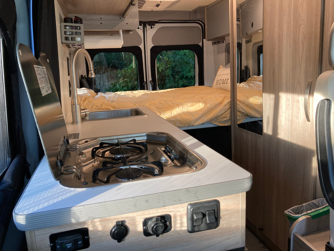 Bright RV rental interior showing a compact kitchen with stove and sink, plus a cozy bed with yellow bedding in the background.