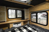 This RV rental interior features a cozy bed, AC unit, shelving, and two windows offering views of a wooded campsite.