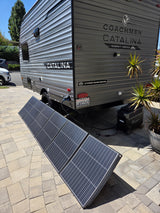 A gray Coachmen Catalina RV rental uses portable solar panels to charge a FOSSIBOT power station on a paved driveway.