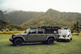 A gray Jeep Gladiator pickup and a white van RV, sticker-covered, parked in a grassy field with misty mountains. Ideal for RV rental trips.